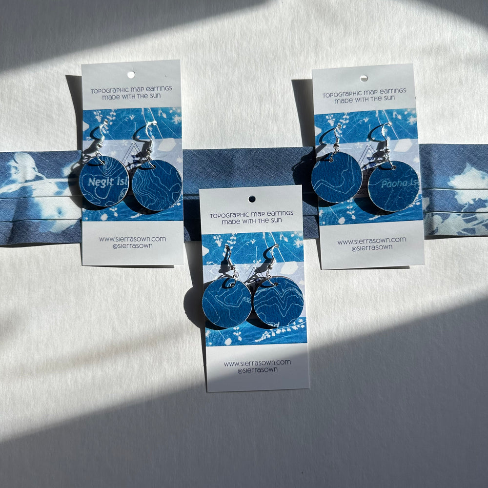 Geographic map earrings on display with branded cards against a light background
