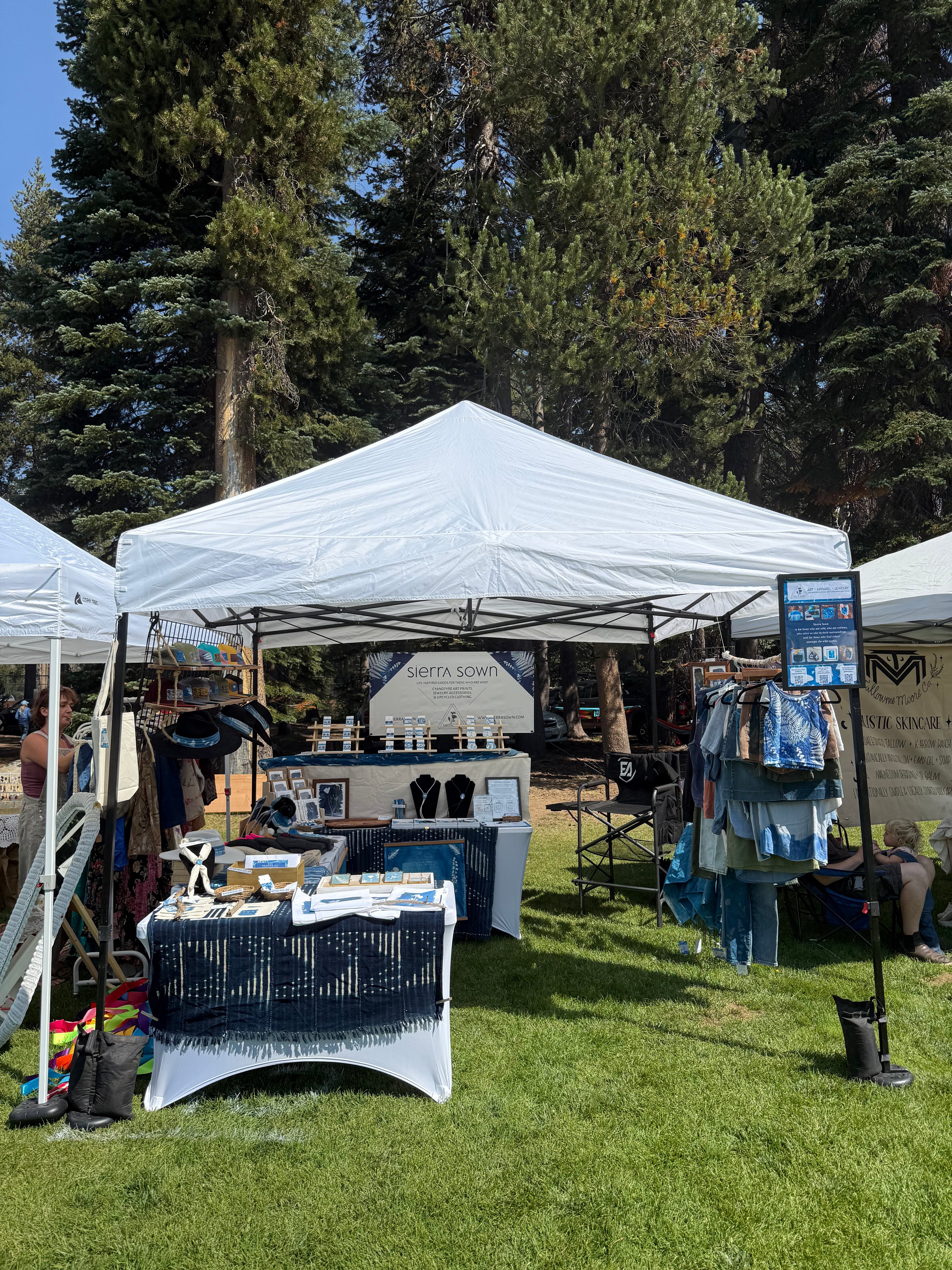 Outdoor event setup with tents, tables, and chairs on a grassy area with trees in the background.