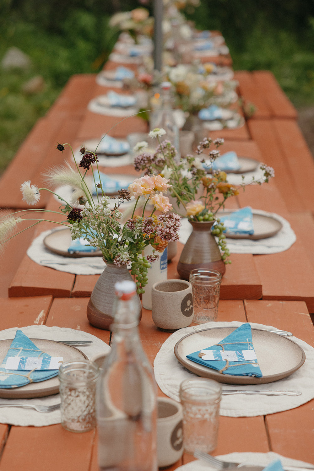 Long outdoor dining table set with place settings, flowers, and drinks on a wooden deck.